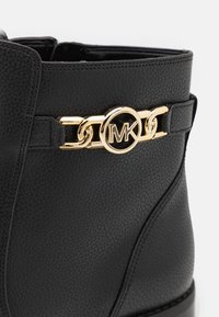 Bottine en cuir noir avec un accent en chaîne dorée et un logo circulaire. La surface texturée comporte des détails de couture.