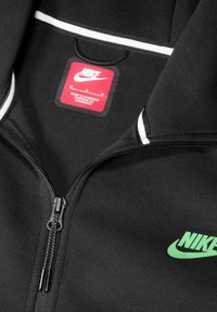 Chaqueta negra con cremallera de Nike que presenta un logo verde, acentos con rayas blancas, tela suave y una etiqueta roja con información de producción.