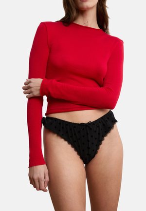 Donna che indossa una maglia rossa a maniche lunghe e biancheria intima nera con pois e rifiniture in pizzo, in piedi davanti a uno sfondo bianco.