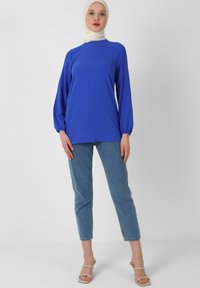 Maglietta blu a maniche lunghe con polsini arricciati, realizzata in tessuto morbido. Abbinata a jeans blu chiaro e sandali beige con cinturini.