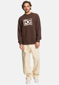 Bruine sweatshirts gemaakt van zachte stof, voorzien van een wit DC Shoes-logo en geribbelde manchetten. Gecombineerd met loszittende crèmekleurige broeken en witte schoenen.