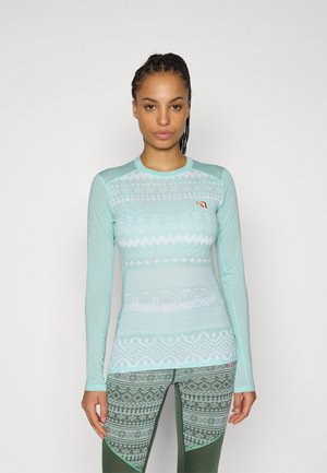 Femme aux cheveux tressés portant un haut à manches longues bleu clair à motifs et un legging vert à motifs, debout devant un fond uni.