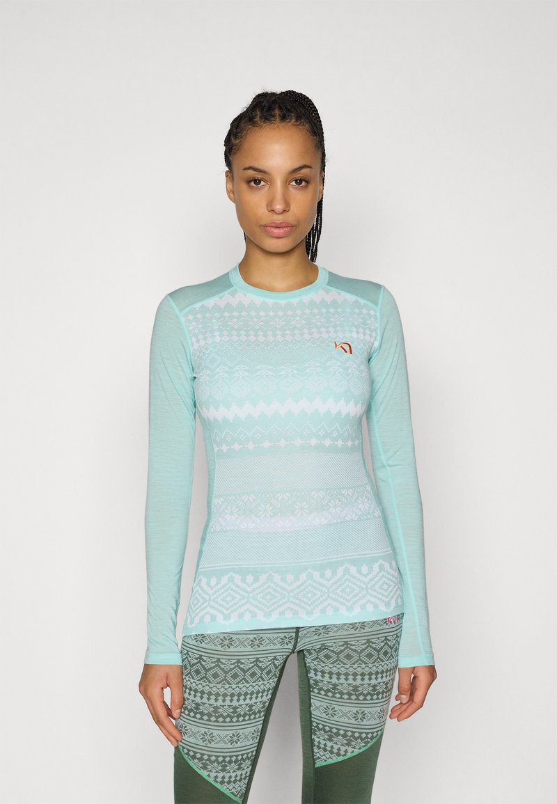 Femme aux cheveux tressés portant un haut à manches longues bleu clair à motifs et un legging vert à motifs, debout devant un fond uni.