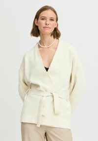 Beige színű, puha anyagból készült kardigán, mély V-nyakkal, széles ujjakkal és anyagból készült övvel. Egyszerű design, minta vagy díszítés nélkül.