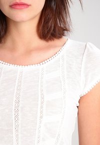 Bílý krátký rukáv top s texturovaným vzorem, který má krajkovou výzdobu podél výstřihu a rukávů, vyrobený z lehké a měkké tkaniny.