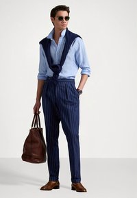 Navy pinstriped trousers, light blue collared shirt, navy sweater draped over shoulders, brown leather shoes, brown bag, dark sunglasses.