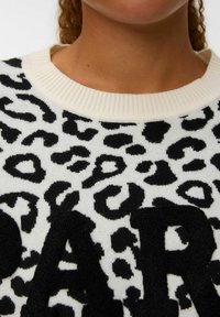 Pull en imprimé léopard noir et blanc avec un col crème côtelé et un détail en lettres noires en peluche. Texture douce, vue rapprochée du tissu.