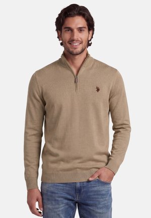 Mann lächelnd, trägt einen beigefarbenen Pullover mit Viertelreißverschluss und kleinem Logo auf der Brust und blaue Jeans, Hand in der Tasche.