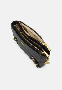 Portefeuille noir texturé avec deux compartiments à fermeture éclair dorés ouverts, doublure en tissu beige, détails en boucle dorée et un charm en forme de logo rond Guess.