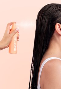 Spray acondicionador sin enjuague IDAWARG en un bote de color melocotón con tapa texturizada, aplicándose sobre cabello lacio y mojado; fondo de color rosa pálido.
