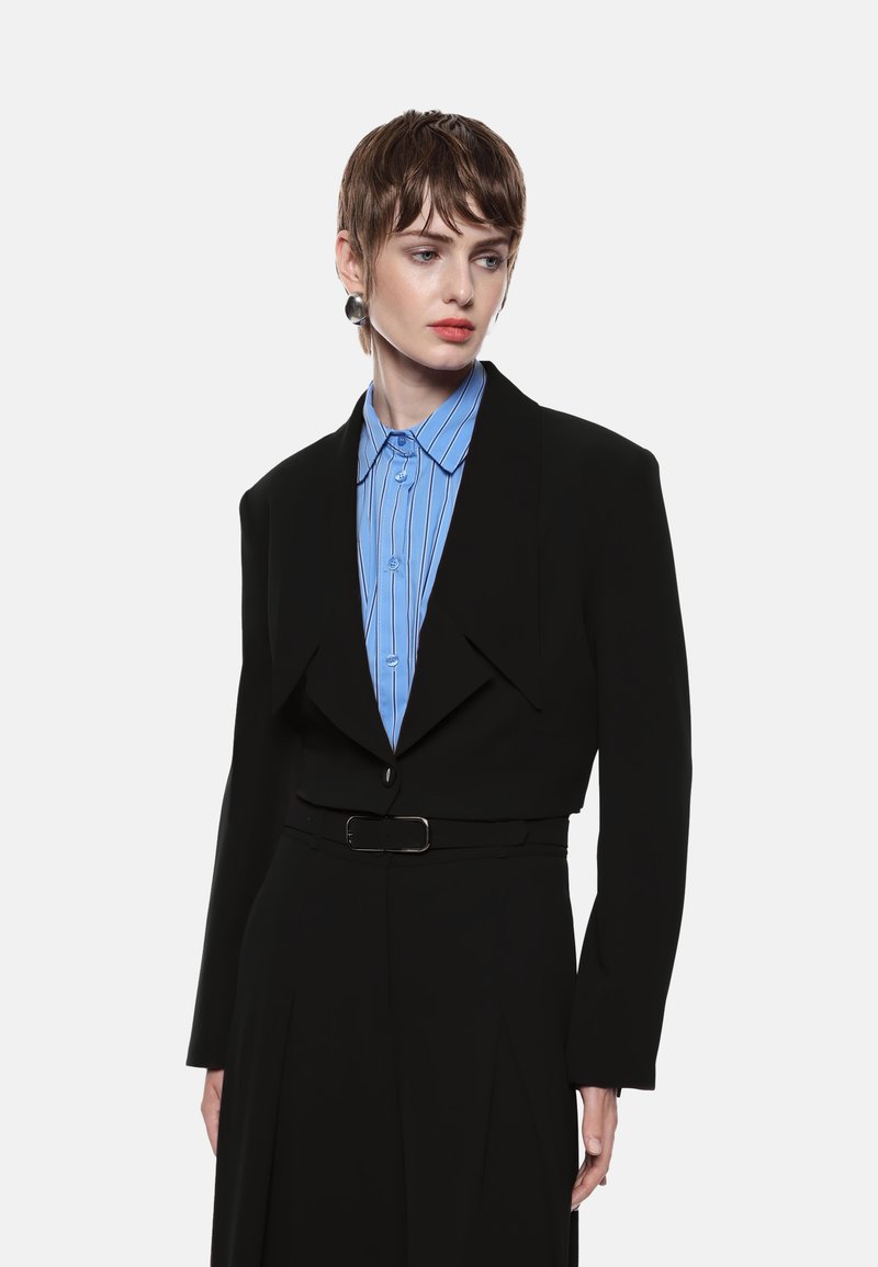 Blazer corto nero con rever affilati sopra una camicia a righe azzurre. Presenta una cintura con una fibbia argentata e pantaloni neri a pieghe dritti.