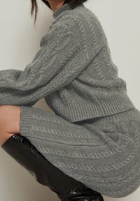 Pull gris en tricot et jupe assortie avec motifs torsadés, col montant et poignets côtelés, associés à des cuissardes noires brillantes.