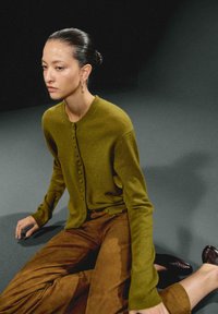 Olivengrøn cardigan med knapdetaljer, kombineret med brune ruskindsbukser. Tekstureret stof og grundlæggende rund halsudskæring. Mørk baggrund.