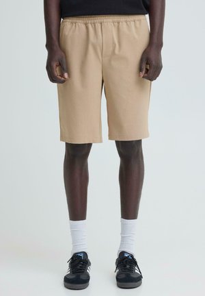Mann trägt beige kniehohe Shorts, weiße Socken und schwarze Turnschuhe, steht vor einem schlichten hellen Hintergrund.