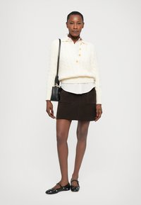 Femme portant un pull en tricot blanc, une chemise blanche à col, une jupe marron foncé avec des poches, des ballerines noires à clous et un sac à bandoulière noir.