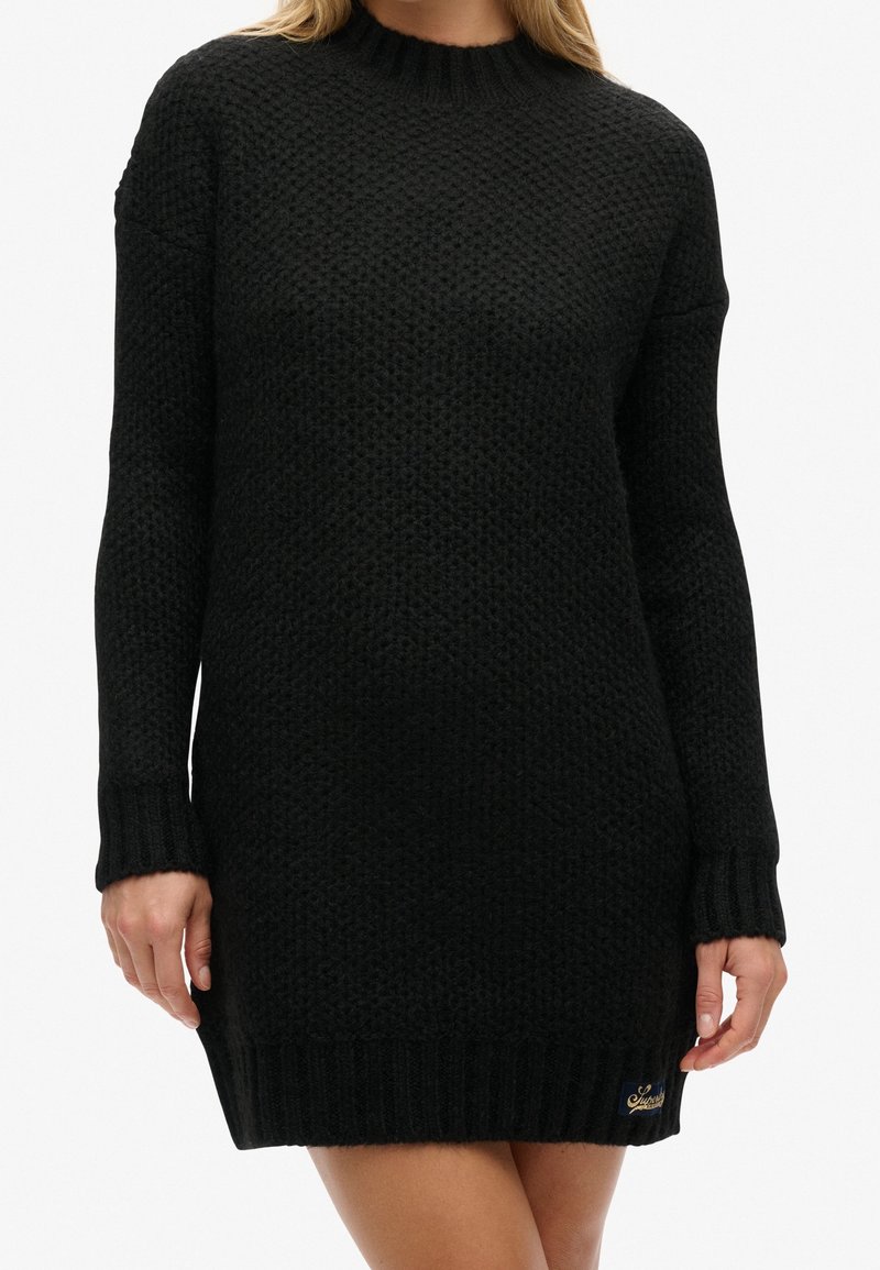 Femme portant une robe pull longue noire en tricot avec un motif texturé et des poignets, ourlet et encolure côtelés.
