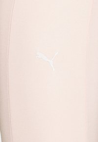 Legginsy Puma w jasnoróżowym kolorze, charakteryzujące się gładką teksturą, bliskim dopasowaniem i białym, ikonicznym logo Puma po lewej stronie. Widoczne detale szwów.
