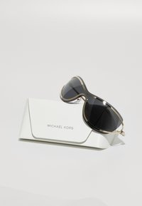 Zwarte zonnebril met een gouden metalen frame en gebogen ontwerp, geplaatst op een witte gestructureerde hoes met de naam "Michael Kors."