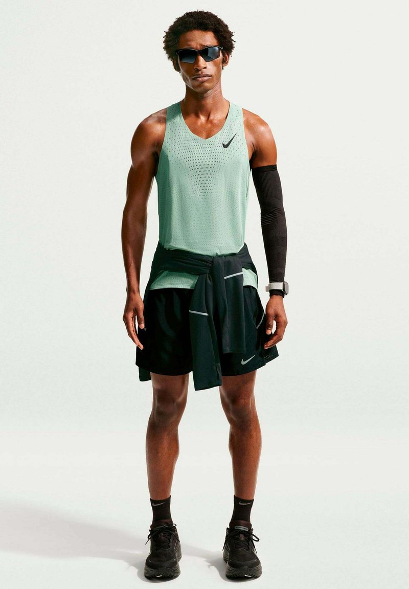 Homme athlétique portant des lunettes de soleil, un débardeur vert menthe, un short noir, une manche noire, une montre connectée et des chaussures de course noires, se tenant face à l’avant.