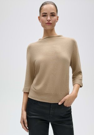 Vrouw met haar naar achteren gebonden, draagt een beige top met lange mouwen en een zwarte broek, staand met één hand in de zak tegen een effen achtergrond.