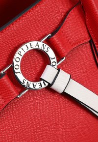 Dettaglio di una borsa a tracolla rossa con texture, caratterizzata da un anello circolare in argento con logo incassato, completata da una tracolla bianca a contrasto con accenti neri.