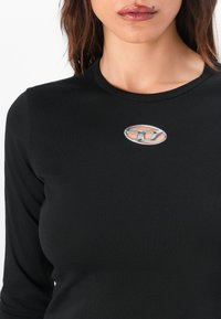 Black long-sleeve top with a fitted design and ribbed texture. Features a silver oval logo accent at the chest.
