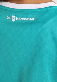Turkoosi urheilupaita, jossa on valkoinen kaulus, takana "DIE MANNSCHAFT" -teksti ja emblem. Valmistettu hengittävästä, teksturoidusta kankaasta.