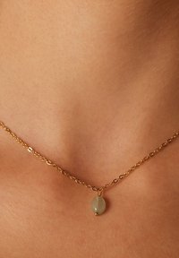 Collar de cadena de oro con un colgante ovalado de color verde pálido y un pequeño acento dorado, reposando sobre un tono de piel claro.