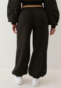 Cilvēks, kurš valkā melnas platās bikses ar aizmugurējām kabatām un elastīgiem piedurkņu galiem, apvienots ar baltām sporta apaviem, stāv pretī vienkāršai fona.