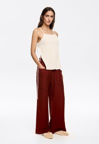 Beige ribbed tank top with side slits paired with wide-leg, dark red pants featuring drawstrings and front pockets. Soft, textured footwear.