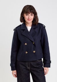 Veste bleu marine à double boutonnage avec des boutons dorés, col structuré et épaulettes, fabriquée dans un tissu texturé.