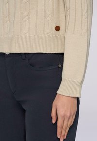 Kremowy sweter w splot warkoczowy z prążkowanymi mankietami, w połączeniu z ciemnoniebieskimi spodniami slim-fit z kieszenią z przodu i metalowym detalem guzika.