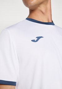 White athletic shirt with a navy ribbed collar and small navy logo on the chest. Made of smooth, lightweight fabric, designed for comfort.