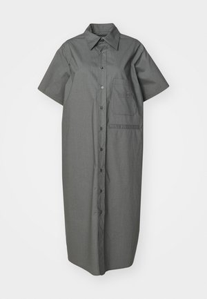 Henkilö pukeutunut harmaaseen lyhythihaiseen nappilista midi-mekkoon, pitää kädessään mustaa pehmeää clutch-laukkua ja käyttää mustia Mary Jane -kenkiä, joissa on valkoiset logohihnat.