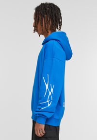 Hoodie albastru cu un buzunar frontal și un design grafic alb pe mâneca stângă. Fabricat dintr-un material moale, cu glugă și croială lejeră.