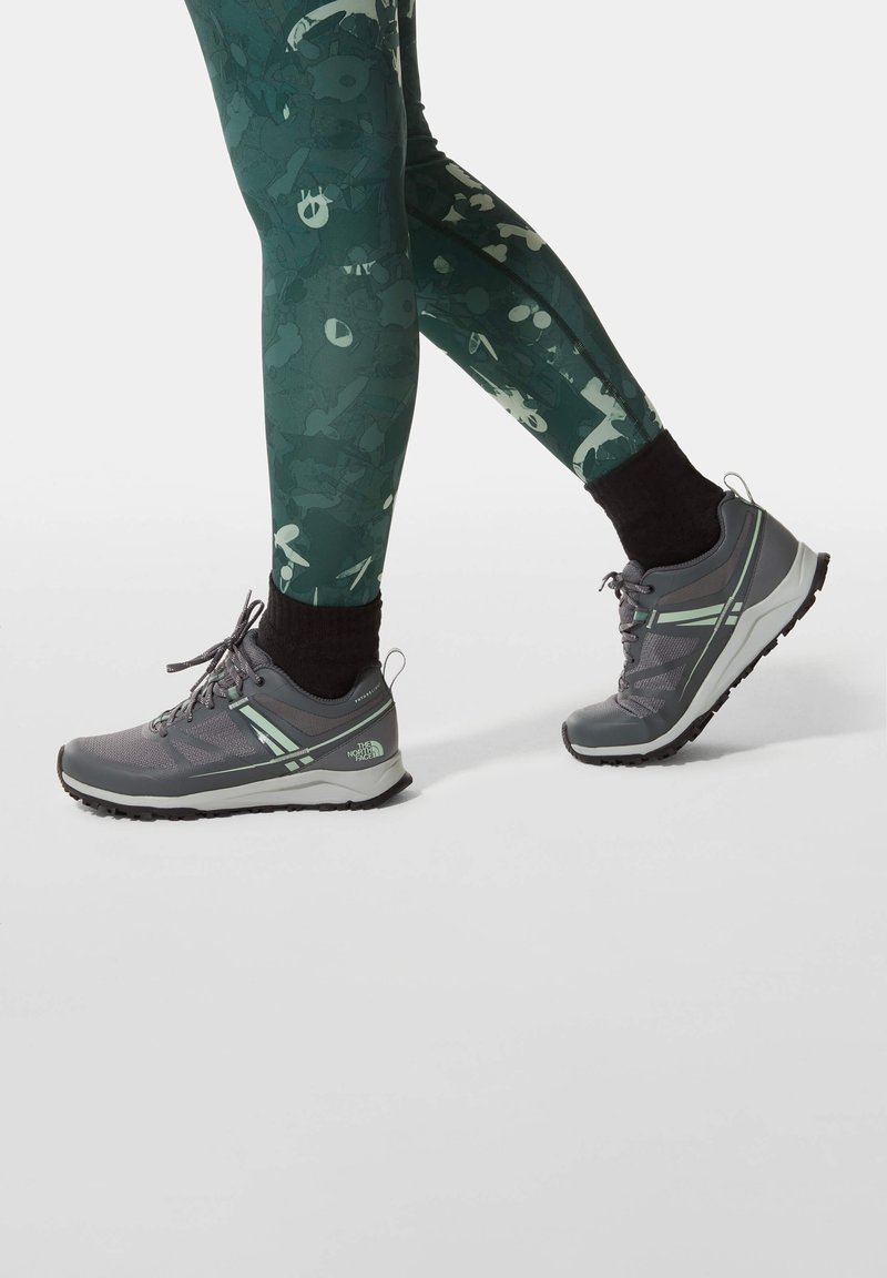 Jambes portant des leggings verts à motifs et des chaussettes noires, avec des chaussures de trail à lacets grises et vert clair en mouvement de marche.
