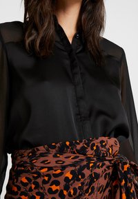 Femme portant un chemisier noir transparent à manches longues rentré dans une jupe léopard brun et orange, nouée à la taille.