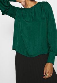 Blusa verde, ampia e morbida, realizzata in materiale soffice. Presenta un collo rotondo, maniche lunghe a sbuffo e dettagli arricciati sulle spalle.