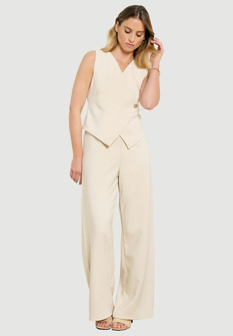 Donna che indossa un gilet asimmetrico senza maniche color crema con scollo a V e pantaloni a gamba larga, che sistema i capelli, su sfondo chiaro uniforme.
