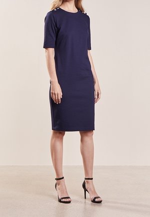 Femme portant une robe bleu marine mi-longue avec manches courtes et détails de boutons aux épaules, associée à des talons hauts noirs à bride cheville.