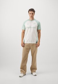 Maglietta da calcio a maniche corte verde chiaro e bianca con un motivo a diamante texturizzato e logo. Il modello indossa pantaloni in corduroy beige e sneakers bianche.