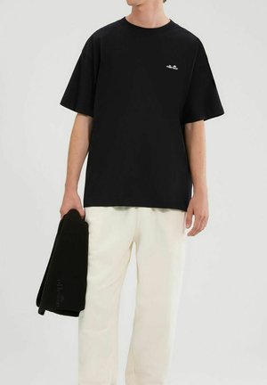 Hombre con camiseta negra de ellesse y pantalones blancos, sosteniendo una prenda negra doblada en la mano izquierda sobre un fondo liso.