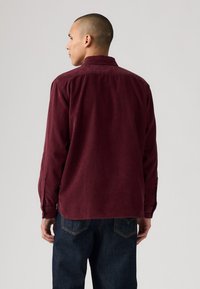 Burgunderfarbendes Cordhemd mit Kragen, langen Ärmeln und geradem Saum. Verfügt über geknöpfte Manschetten und eine subtile Textur. Getragen mit dunkelblauen Jeans.