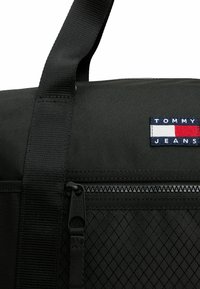 Borsa a tracolla nera con superficie testurizzata, dotata di una tasca con zip, una spallina spessa e una distintiva toppa con logo in rosso, bianco e blu.