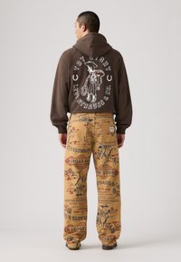Sudadera marrón con un gráfico blanco de "Toy Story" en la parte posterior; pantalones amarillos con un estampado colorido de personajes de dibujos animados y texto.