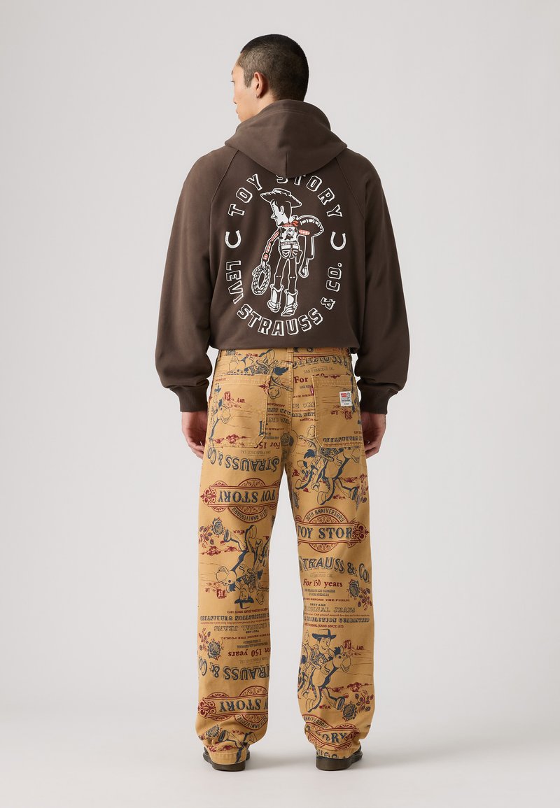 Brun hoodie med vit "Toy Story" grafik på ryggen; gula byxor med färgglad tryck av tecknade karaktärer och text.
