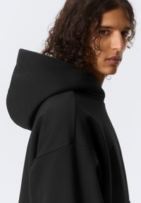 Svart hettegenser i mykt materiale. Har en avslappet passform med hette som er høy og strukturert, og glatt tekstur.