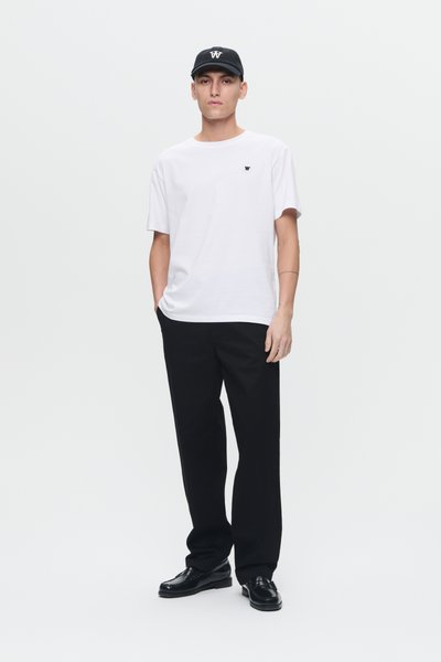 Jonge man die staat en een wit T-shirt met een klein logo draagt, zwarte broek, zwarte schoenen en een zwarte pet met een wit logo.