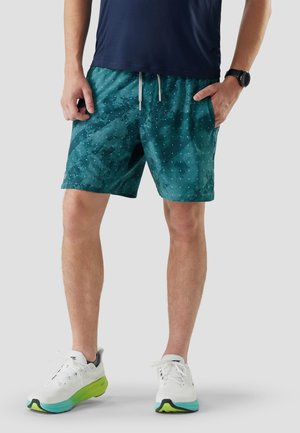Homme portant un short à motifs verts d'eau, des baskets blanches avec des semelles vertes, une chemise bleu marine et une montre intelligente noire, debout les mains dans les poches.