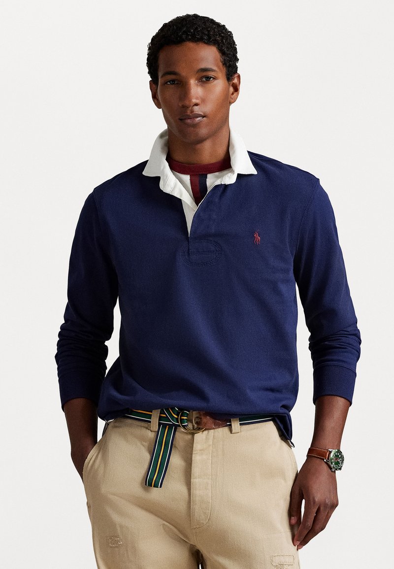 Marineblauw poloshirt met lange mouwen en een witte kraag, voorzien van een klein geborduurd logo. Gecombineerd met kaki broek en een gestreepte riem.
